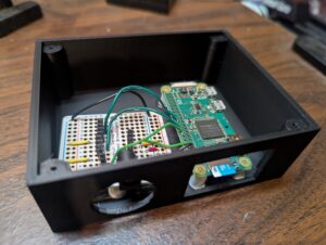 Black project box without the top. It has a large round opening where the barrel jack for power resids, and a large rectangular opening where the micro SC card can be accessed. Inside are the Pi Zero W and the solderable breadboard.